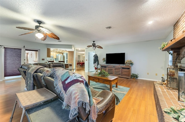 living room featuring ceiling fan, a brick fireplace, a textured ceiling, and light wood-type flooring