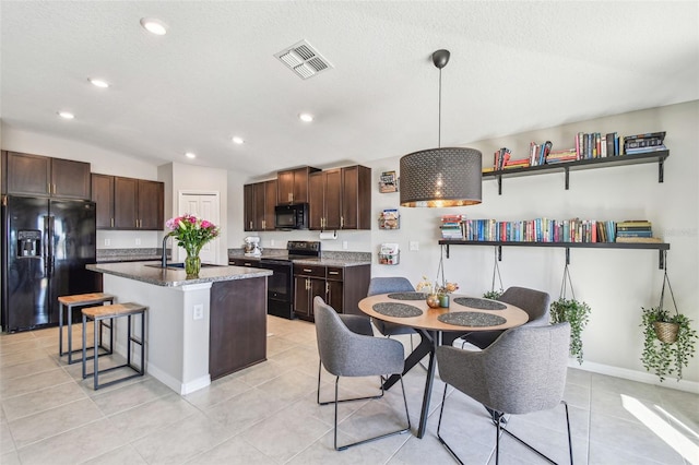 kitchen featuring a breakfast bar, a kitchen island with sink, hanging light fixtures, black appliances, and dark stone counters