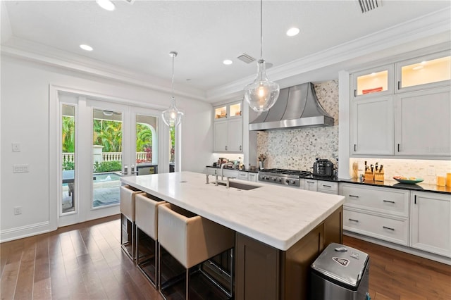 kitchen featuring a sink, wall chimney range hood, an island with sink, dark wood finished floors, and crown molding