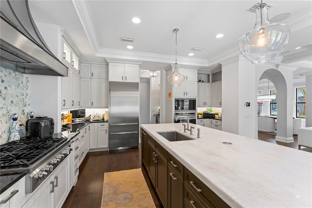 kitchen featuring decorative light fixtures, built in appliances, range hood, crown molding, and a sink