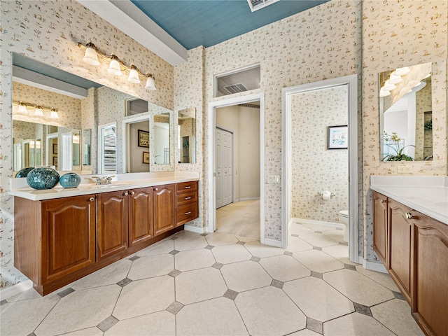 full bathroom featuring wallpapered walls, baseboards, toilet, a sink, and two vanities