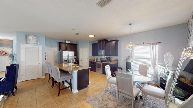 kitchen featuring appliances with stainless steel finishes, a kitchen island with sink, dark brown cabinetry, light tile patterned flooring, and decorative light fixtures