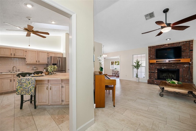 kitchen with stainless steel refrigerator with ice dispenser, sink, tasteful backsplash, a brick fireplace, and light brown cabinets