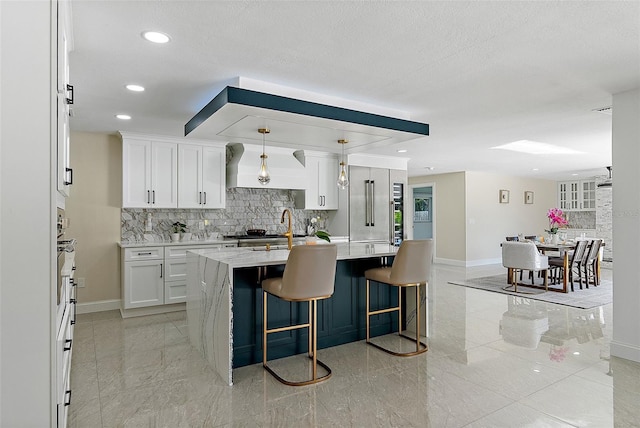 kitchen featuring premium range hood, a kitchen island with sink, a kitchen breakfast bar, white cabinets, and decorative light fixtures