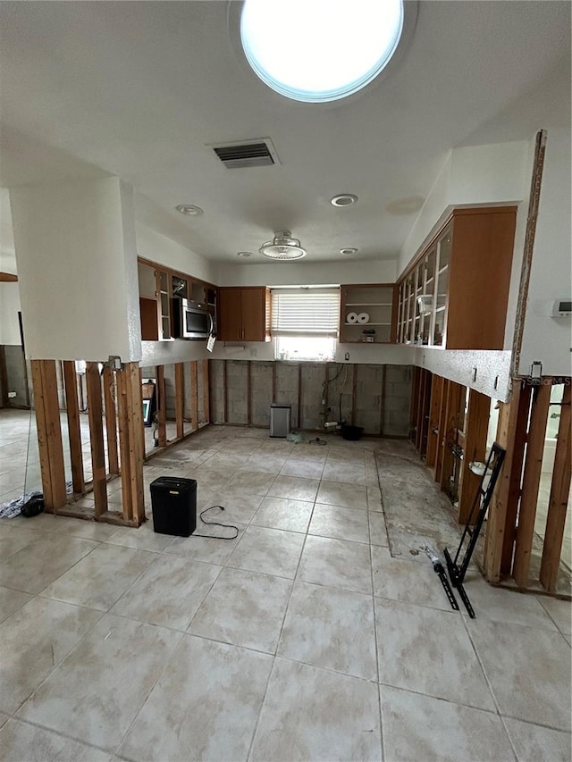 kitchen featuring stainless steel microwave, visible vents, glass insert cabinets, brown cabinetry, and light tile patterned floors