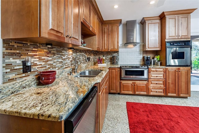 kitchen with wall chimney exhaust hood, brown cabinets, light stone countertops, stainless steel appliances, and a sink