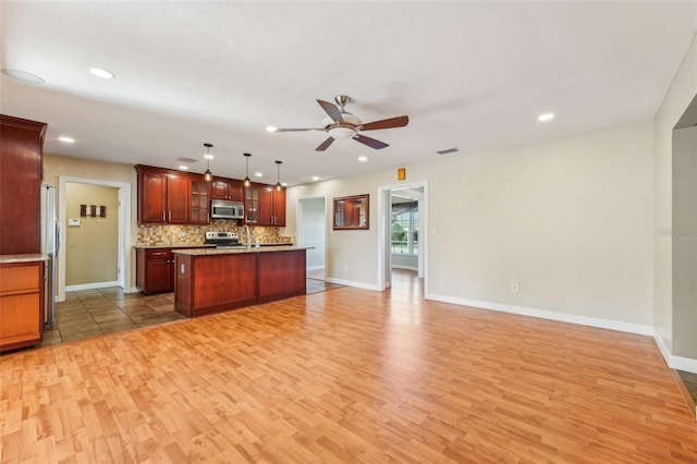 kitchen featuring appliances with stainless steel finishes, decorative light fixtures, an island with sink, wood-type flooring, and backsplash