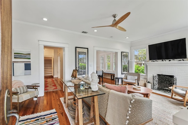 living room featuring a brick fireplace, hardwood / wood-style flooring, ornamental molding, and ceiling fan