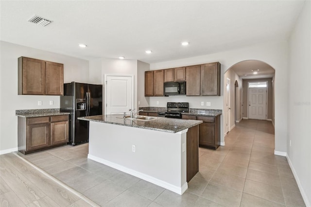 kitchen featuring light tile patterned flooring, sink, dark stone countertops, black appliances, and a center island with sink