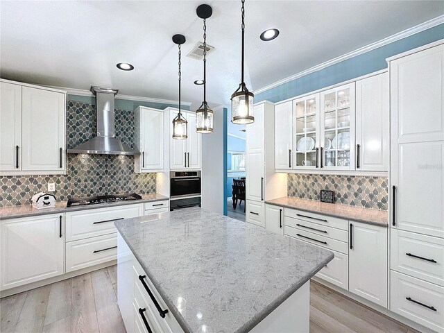 kitchen with white cabinets, a kitchen island, glass insert cabinets, stainless steel appliances, and wall chimney range hood