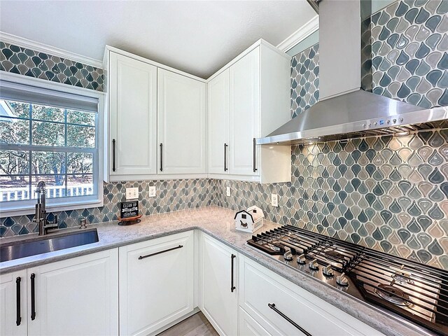 kitchen with a sink, white cabinets, stainless steel gas stovetop, wall chimney exhaust hood, and crown molding