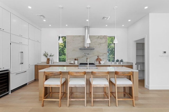 kitchen featuring white cabinetry, a spacious island, wall chimney range hood, paneled built in refrigerator, and pendant lighting