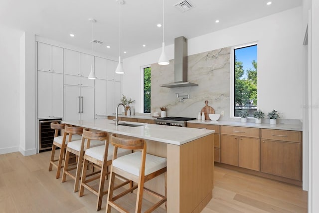 kitchen featuring a center island with sink, hanging light fixtures, wall chimney range hood, sink, and white cabinetry