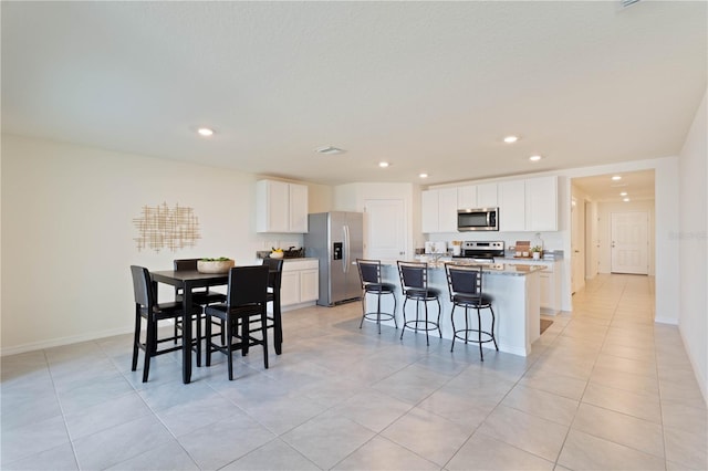 kitchen with light tile patterned floors, a breakfast bar, stainless steel appliances, white cabinets, and a center island with sink