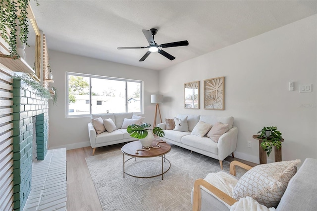 living room featuring ceiling fan, a textured ceiling, wood finished floors, baseboards, and a brick fireplace