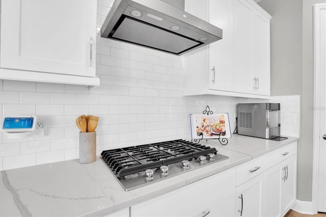 kitchen with stainless steel gas stovetop, white cabinets, backsplash, light stone countertops, and wall chimney range hood