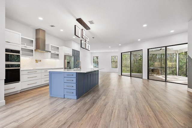 kitchen featuring a kitchen island with sink, double oven, stainless steel refrigerator with ice dispenser, white cabinets, and wall chimney exhaust hood