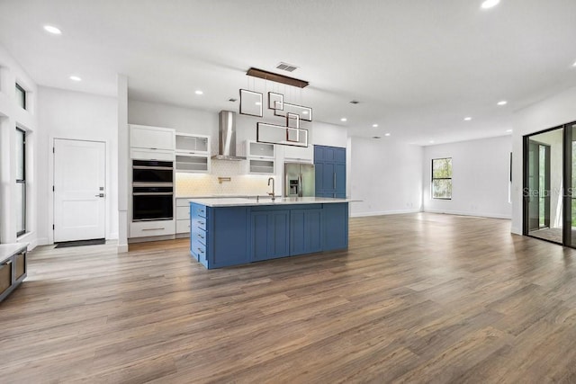 kitchen featuring blue cabinetry, stainless steel fridge with ice dispenser, an island with sink, wall chimney range hood, and white cabinets