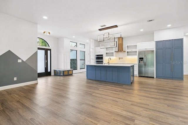 kitchen featuring stainless steel fridge with ice dispenser, white cabinets, wall chimney range hood, a kitchen island with sink, and backsplash