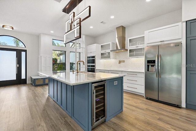 kitchen featuring a kitchen island with sink, stainless steel refrigerator with ice dispenser, white cabinets, beverage cooler, and wall chimney exhaust hood