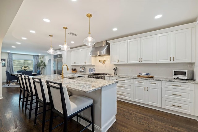 kitchen featuring decorative light fixtures, white cabinetry, sink, wall chimney range hood, and a center island with sink