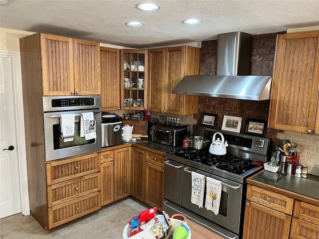 kitchen featuring dark countertops, wall chimney exhaust hood, appliances with stainless steel finishes, brown cabinets, and glass insert cabinets