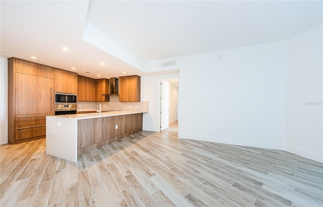 kitchen featuring black microwave, oven, kitchen peninsula, wall chimney range hood, and light hardwood / wood-style flooring