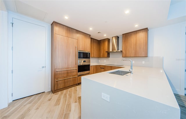 kitchen with black appliances, sink, kitchen peninsula, wall chimney range hood, and light hardwood / wood-style flooring