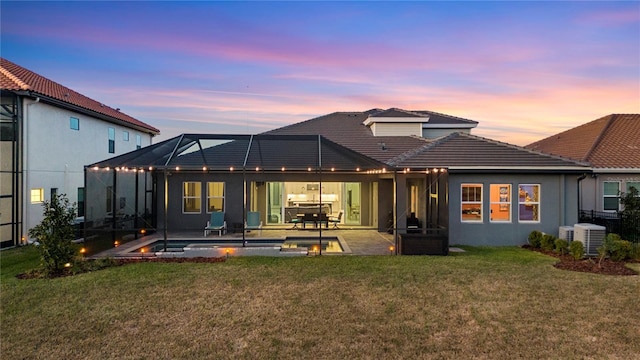 back house at dusk with cooling unit, a patio area, glass enclosure, and a lawn