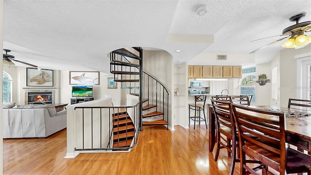 dining area with ceiling fan, a textured ceiling, light wood-style flooring, a glass covered fireplace, and stairs