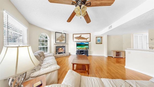 living area with a textured ceiling, light wood-type flooring, a fireplace, ceiling fan, and baseboards