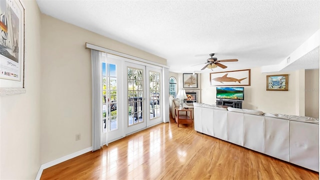 living area with light wood-style floors, a textured ceiling, a glass covered fireplace, ceiling fan, and baseboards