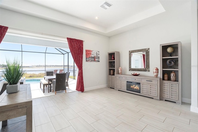 interior space featuring baseboards, visible vents, a tray ceiling, a water view, and a glass covered fireplace