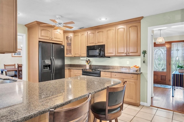 kitchen with light tile patterned floors, black appliances, a kitchen bar, light brown cabinetry, and dark stone counters