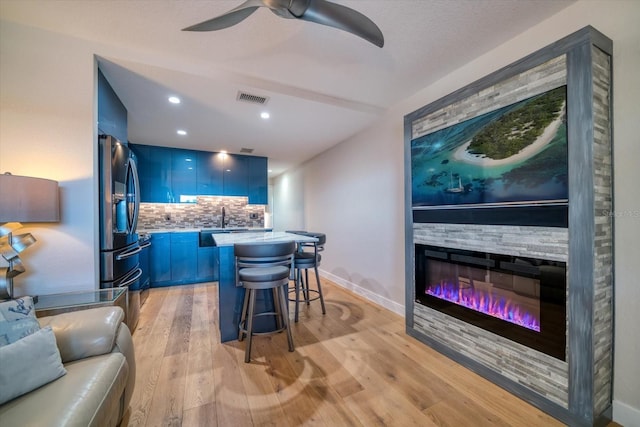 bar with a stone fireplace, blue cabinets, backsplash, light hardwood / wood-style floors, and stainless steel refrigerator with ice dispenser