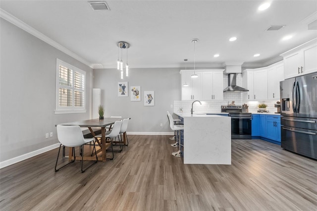 kitchen featuring blue cabinets, white cabinetry, hanging light fixtures, stainless steel appliances, and wall chimney range hood