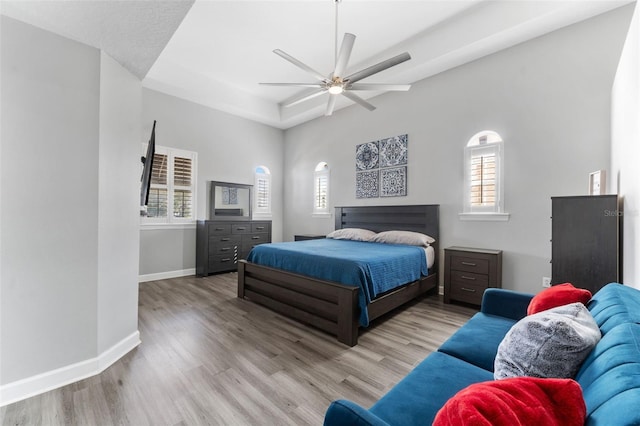 bedroom with ceiling fan, a raised ceiling, multiple windows, and light wood-type flooring