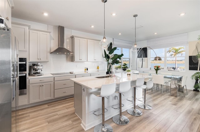 kitchen with wall chimney range hood, a breakfast bar area, hanging light fixtures, stainless steel appliances, and a center island with sink