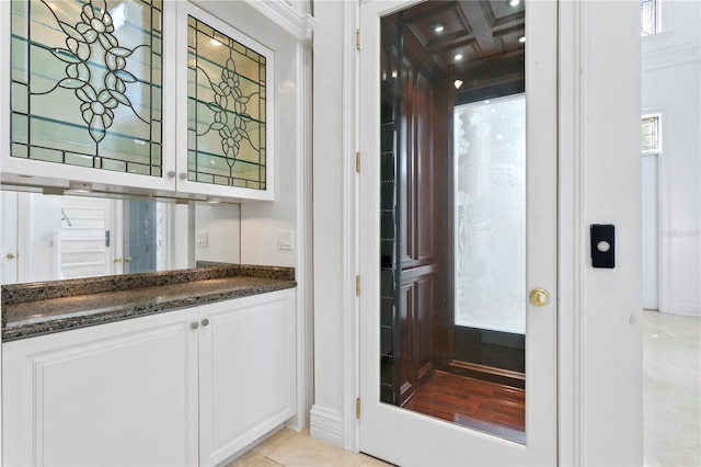 bar with dark stone countertops, coffered ceiling, ornamental molding, white cabinets, and light tile patterned flooring