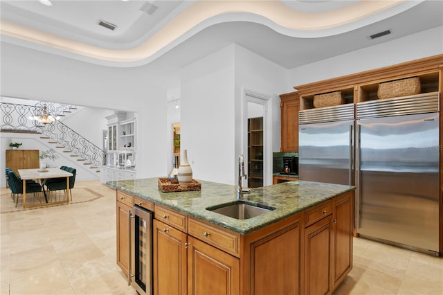 kitchen featuring built in fridge, sink, beverage cooler, a kitchen island with sink, and light stone counters