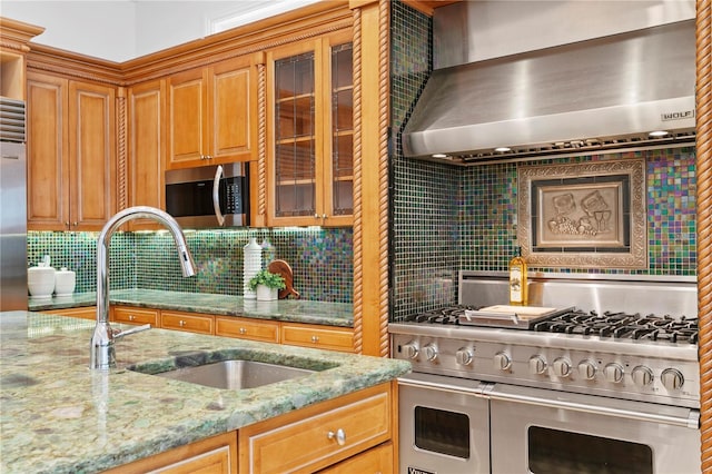 kitchen featuring high end appliances, wall chimney range hood, light stone counters, and sink