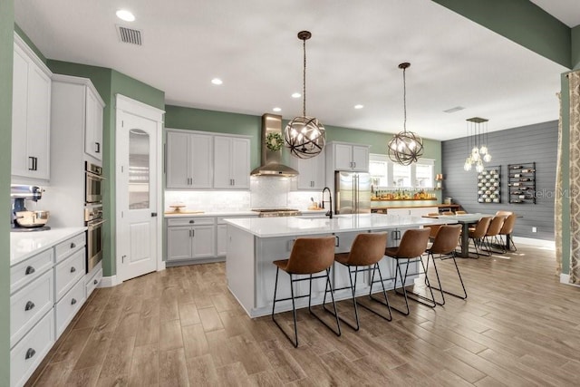 kitchen featuring wall chimney range hood, white cabinetry, stainless steel appliances, a center island with sink, and decorative light fixtures