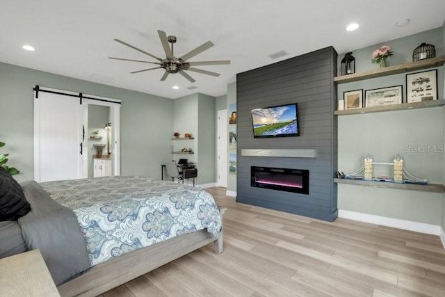 bedroom with ceiling fan, a large fireplace, a barn door, and light hardwood / wood-style floors