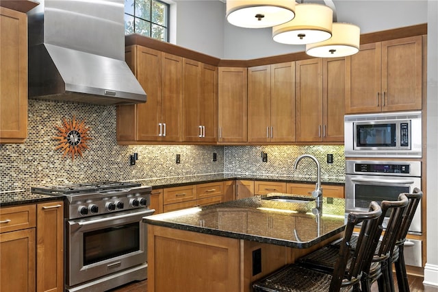 kitchen with wall chimney range hood, sink, appliances with stainless steel finishes, a kitchen island with sink, and a kitchen breakfast bar