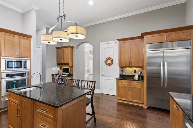 kitchen with a breakfast bar, hanging light fixtures, ornamental molding, a kitchen island with sink, and built in appliances