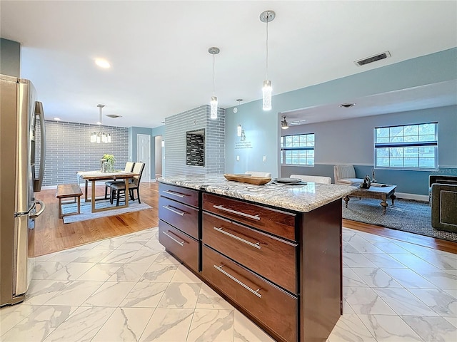 kitchen with a kitchen island, stainless steel fridge, hanging light fixtures, light stone counters, and dark brown cabinetry