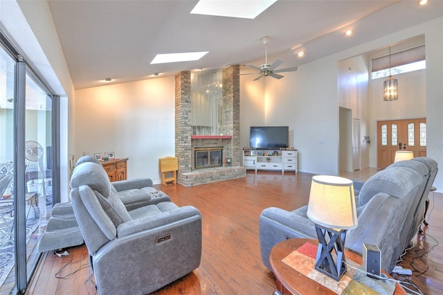 living room with hardwood / wood-style flooring, ceiling fan, a skylight, high vaulted ceiling, and a brick fireplace