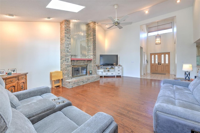 living room featuring high vaulted ceiling, a skylight, ceiling fan, a brick fireplace, and light hardwood / wood-style flooring