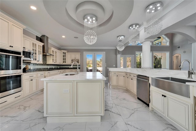 kitchen with cream cabinetry, a tray ceiling, an island with sink, and wall chimney range hood