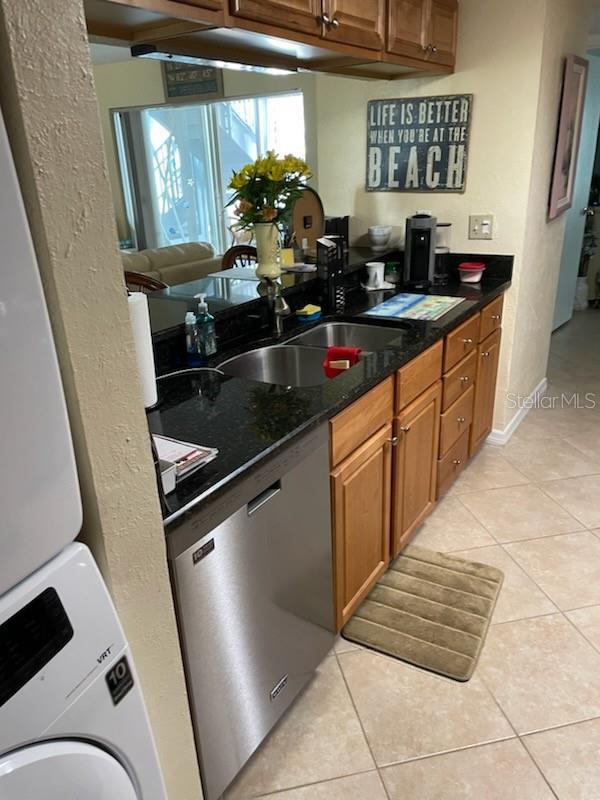 kitchen featuring sink, dark stone countertops, stacked washer / drying machine, stainless steel dishwasher, and light tile patterned floors